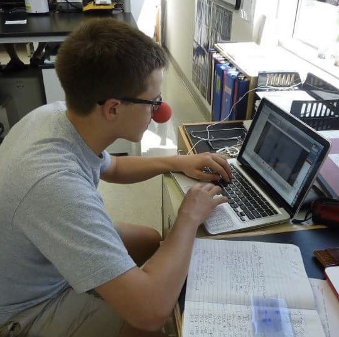 The editor at a laptop wearing a red clown nose, flanked by handwritten notebooks